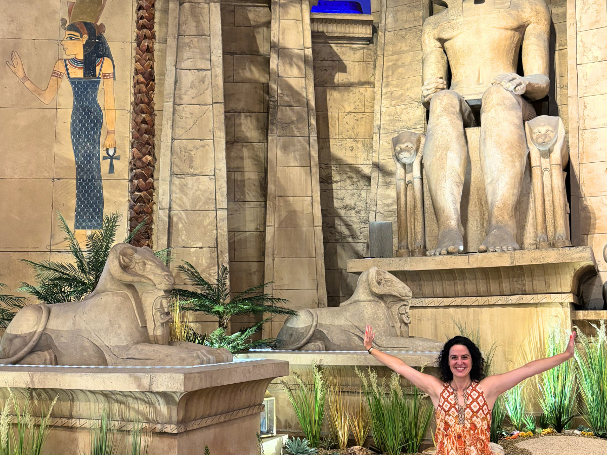 Woman posing with arms raised beside a large Egyptian statue and two sphinxes at an ancient-looking wall scene.