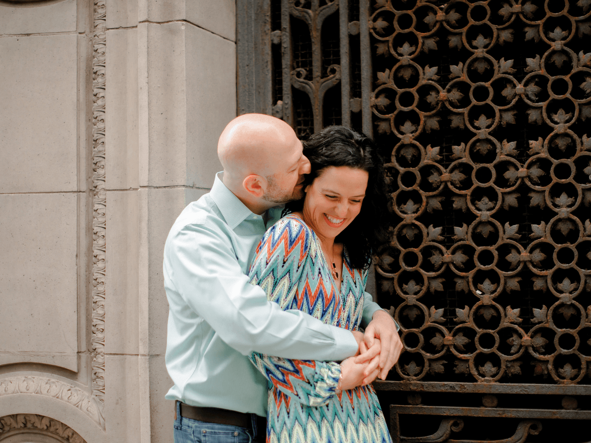 A bald man in a light blue shirt embraces and kisses a smiling woman wearing a turquoise zigzag dress in front of a decorative metal gate.