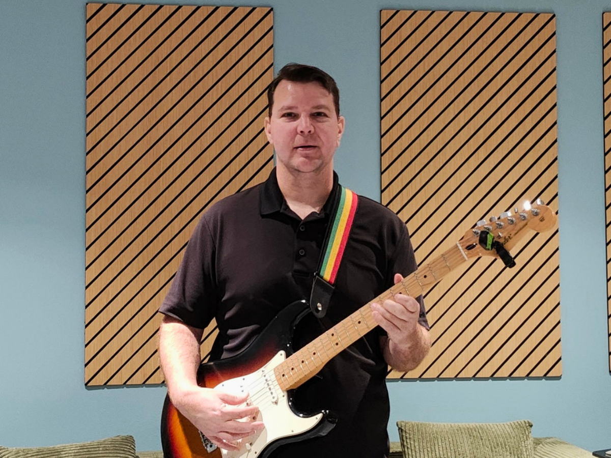 Man wearing a black polo plays a sunburst Fender Stratocaster in a studio with diagonal wooden sound panels on a blue wall behind him, strumming the guitar.