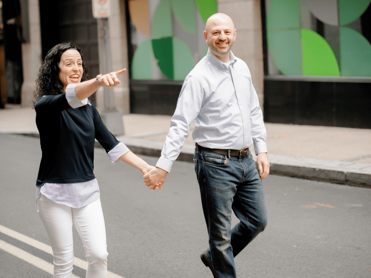 Smiling couple walking hand in hand down a city street, the woman pointing ahead as they travel together.