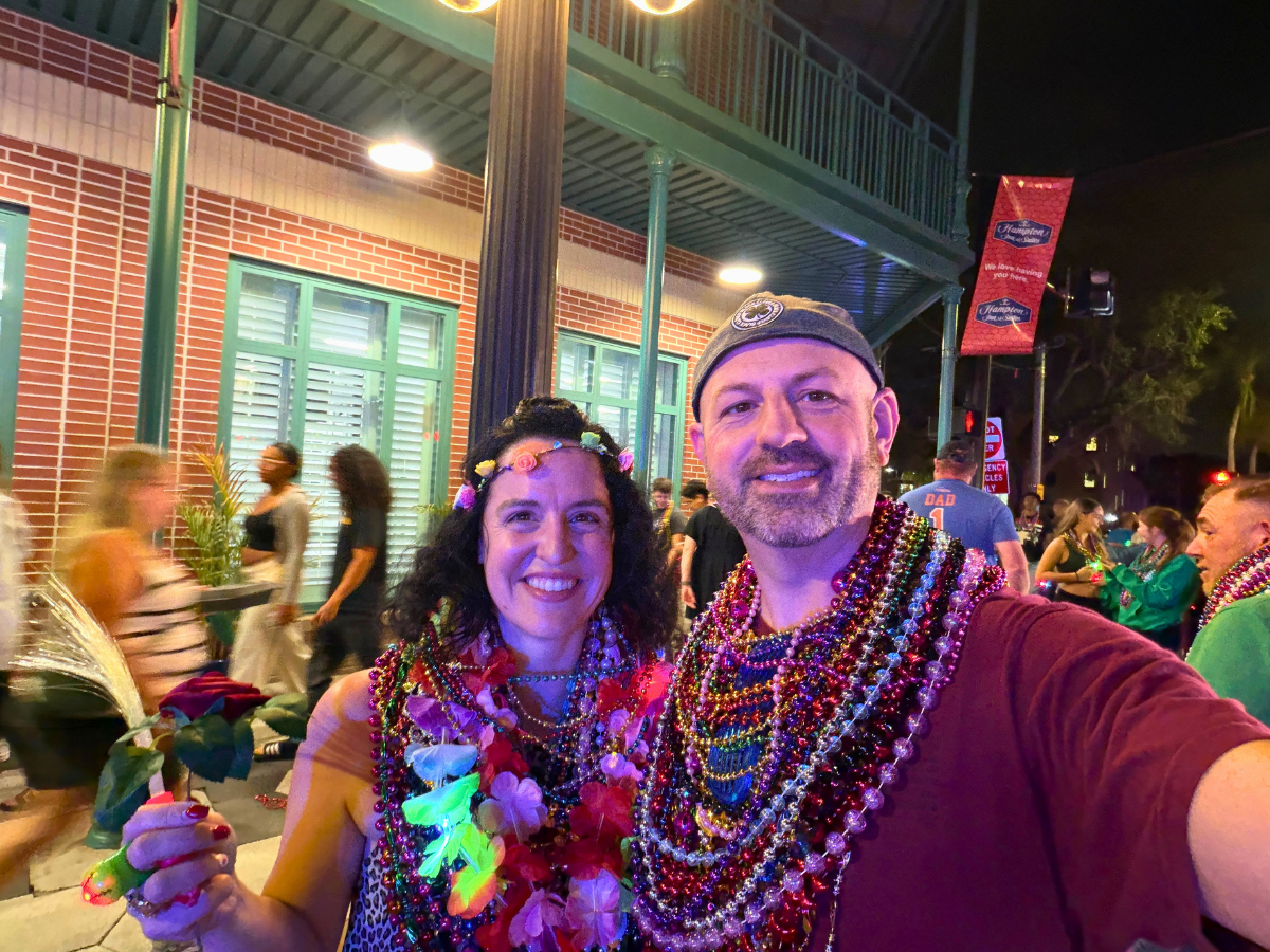 Smiling couple taking a night selfie at a street festival, wearing colorful bead necklaces and leis; woman holds a flower.