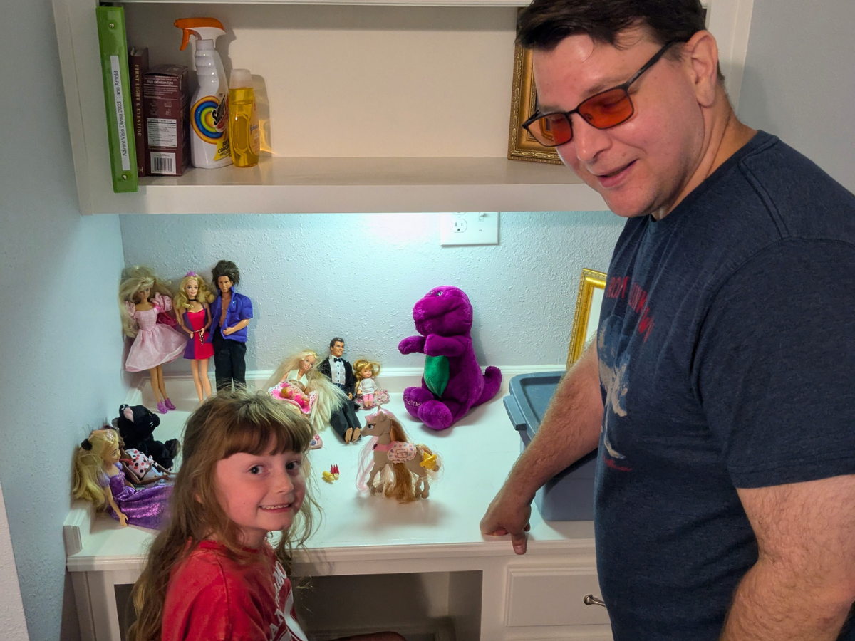 A man wearing orange-tinted glasses and a blue shirt stands beside a young girl as they view a lit cabinet shelf filled with dolls.