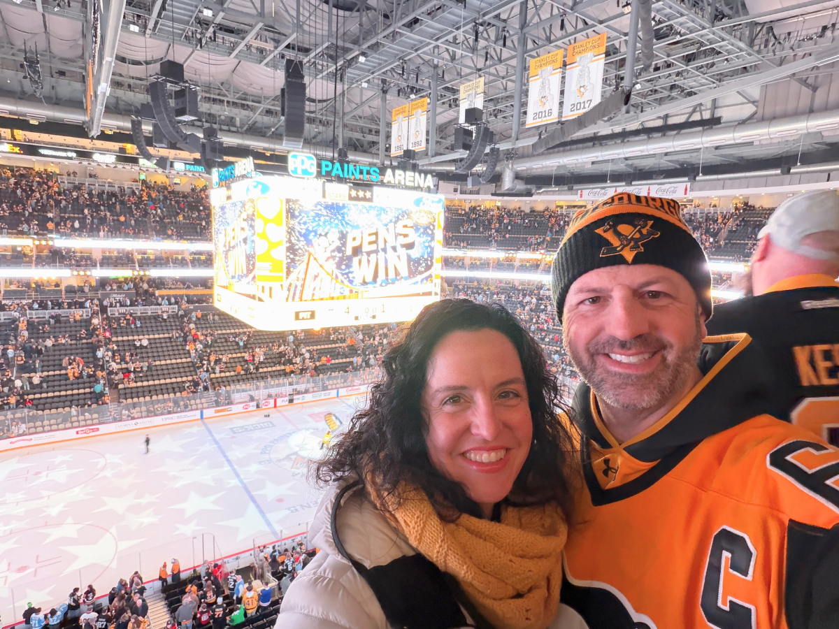 Smiling couple pose for a selfie in an NHL arena with a scoreboard reading 'PENS WIN' and fans in the stands behind them at PPG Paints Arena in Pittsburgh.