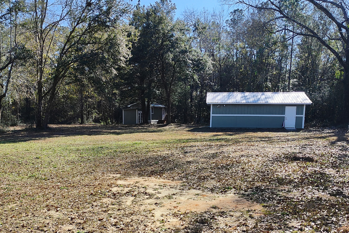 Open yard with two small sheds near a tree line; larger blue shed with white door on the right.