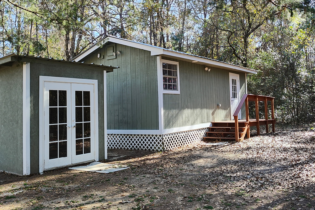 Green wooden cabin with white trim and front steps in a wooded yard, next to a small shed with French doors.