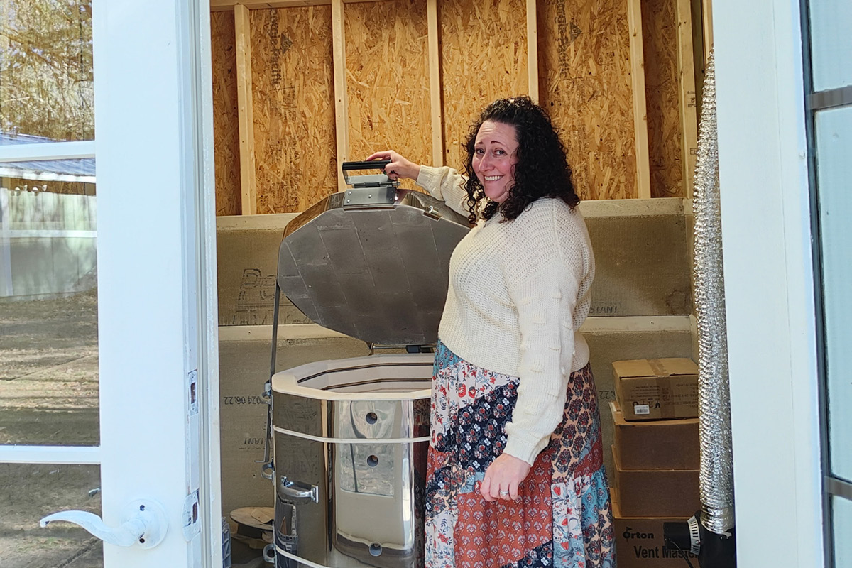 Smiling woman in a cream sweater standing by a large outdoor smoker with its lid open inside a wood-framed workspace.