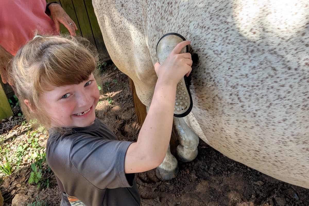 Young girl brushing a horse's hoof with a grooming tool, smiling at the camera outdoors.