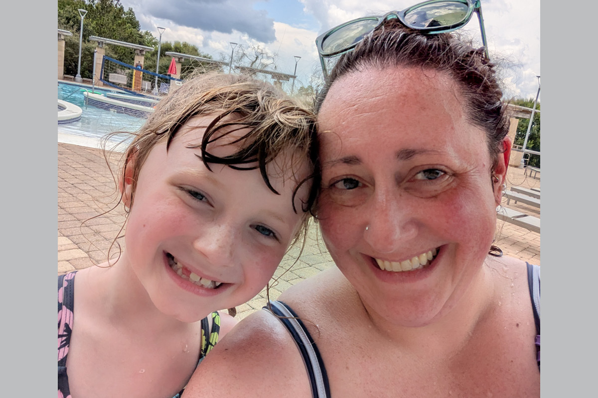 Mother and daughter smiling for a close-up selfie at an outdoor pool on a sunny day, with lounge chairs in the background.