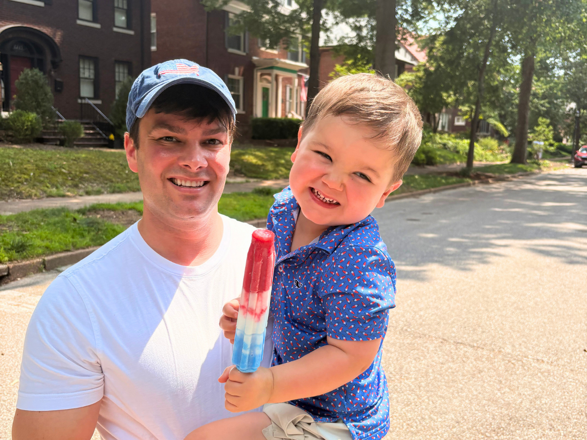 Father and young son pose on a sunny neighborhood street; the boy holds a red, white, and blue popsicle with a big smile.
