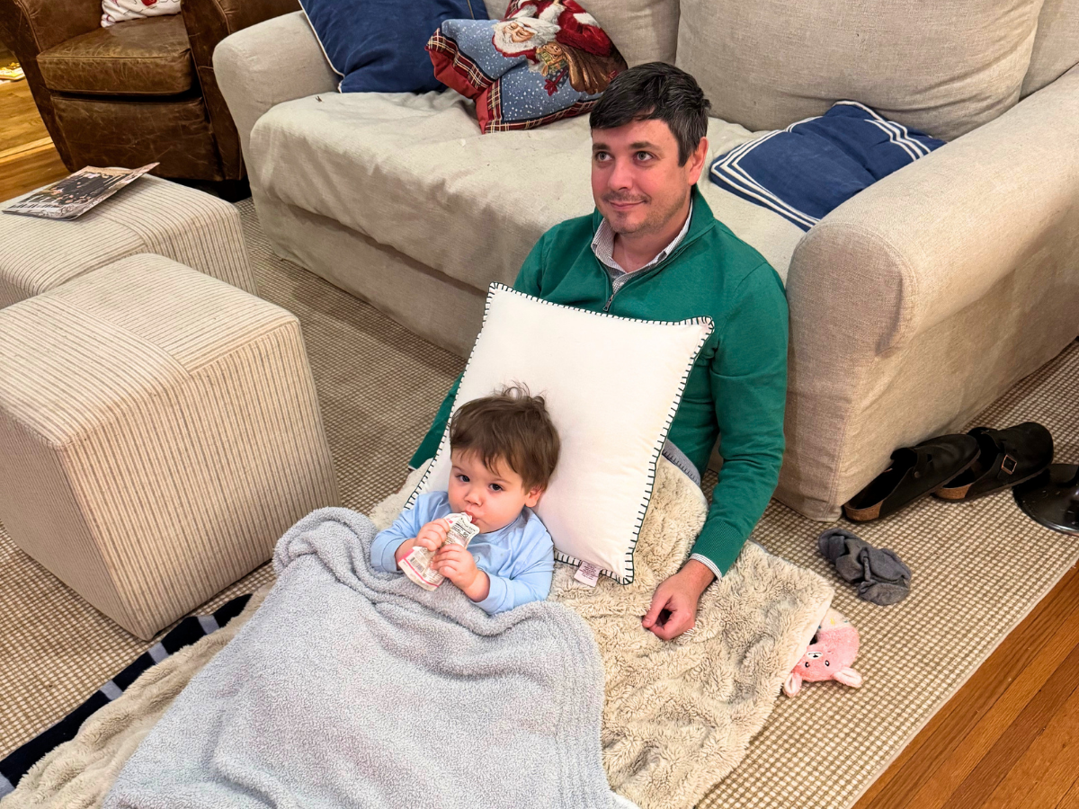 Father sits on the living room floor with a baby wrapped in a blanket and a white pillow behind them, beside a beige sofa.