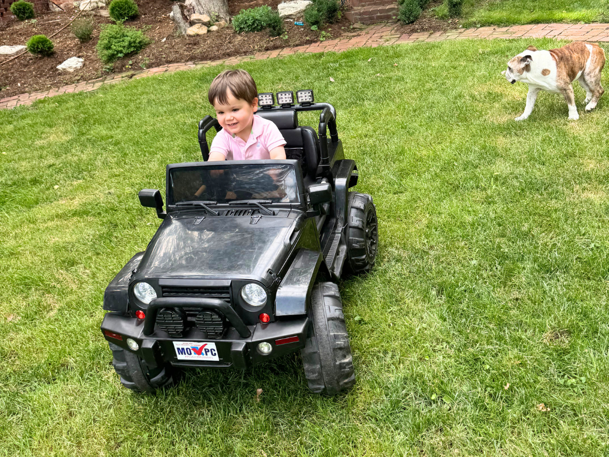 A young child in a pink shirt drives a black toy jeep across a grassy yard, with a bulldog in the background.