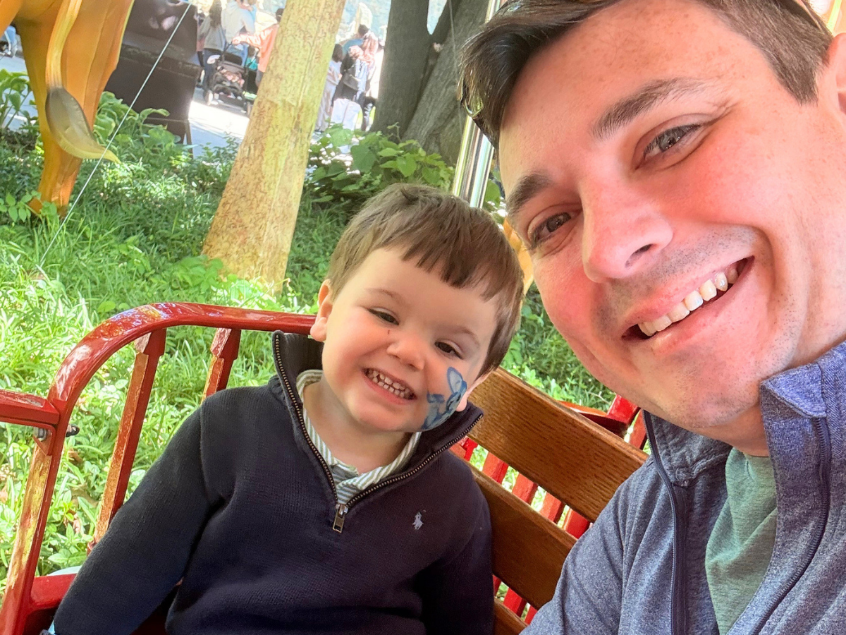 Smiling man and young boy posing for a selfie on a park ride, boy has blue face paint on his cheek.