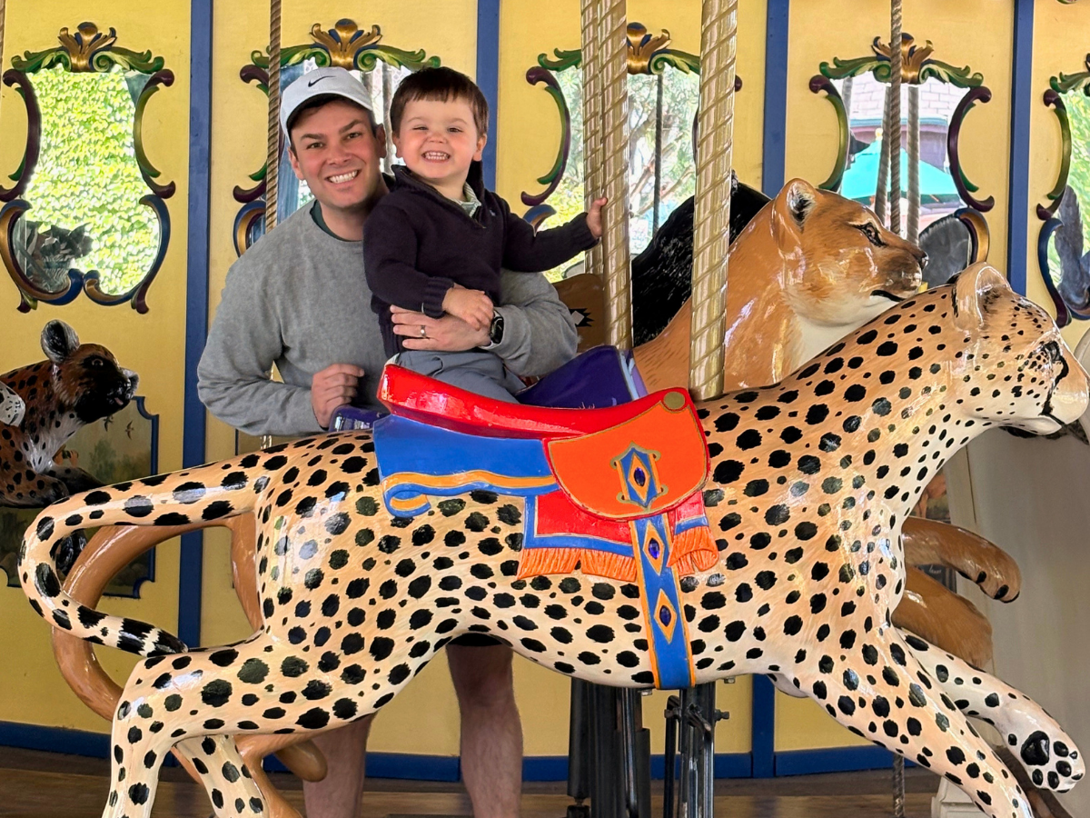 Father holding a smiling toddler while riding a colorful carousel horse with a decorative saddle and yellow backdrop.