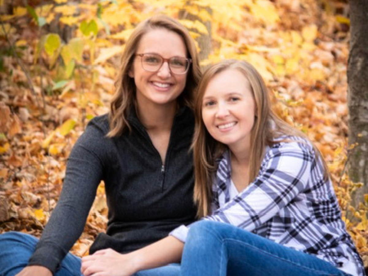 Two women sitting together on autumn leaves, smiling at the camera, wearing casual sweaters.