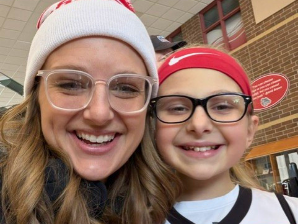 Two smiling people taking a selfie indoors; adult wearing a white knit hat and clear-framed glasses, child in a red headband and black-framed glasses.