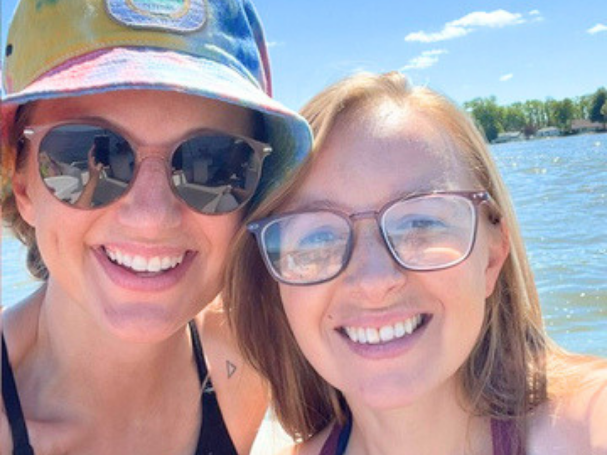 Two women smiling for a selfie on a sunny lakeside, one wearing a colorful cap and sunglasses, the other with glasses beside water.