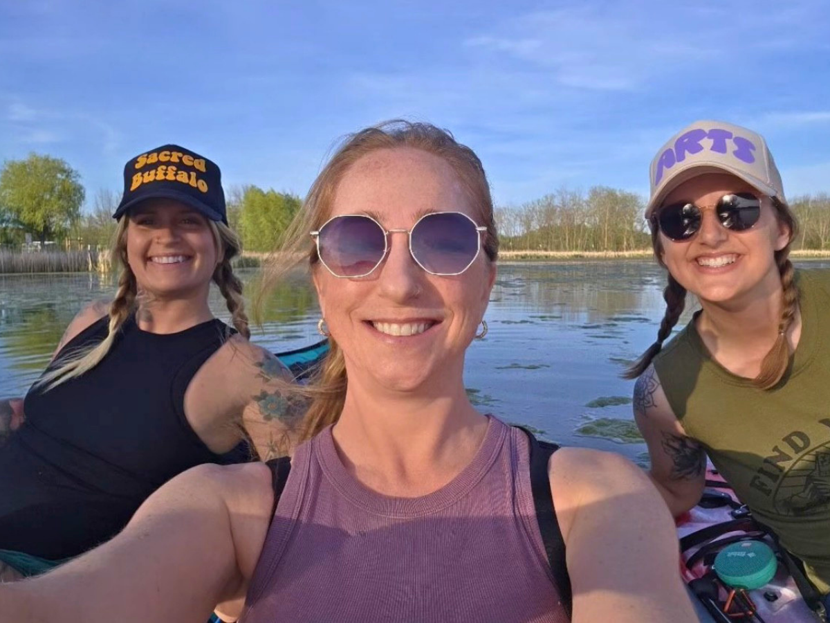 Three smiling friends on a boat taking a selfie on a sunny lake, wearing hats and sunglasses.