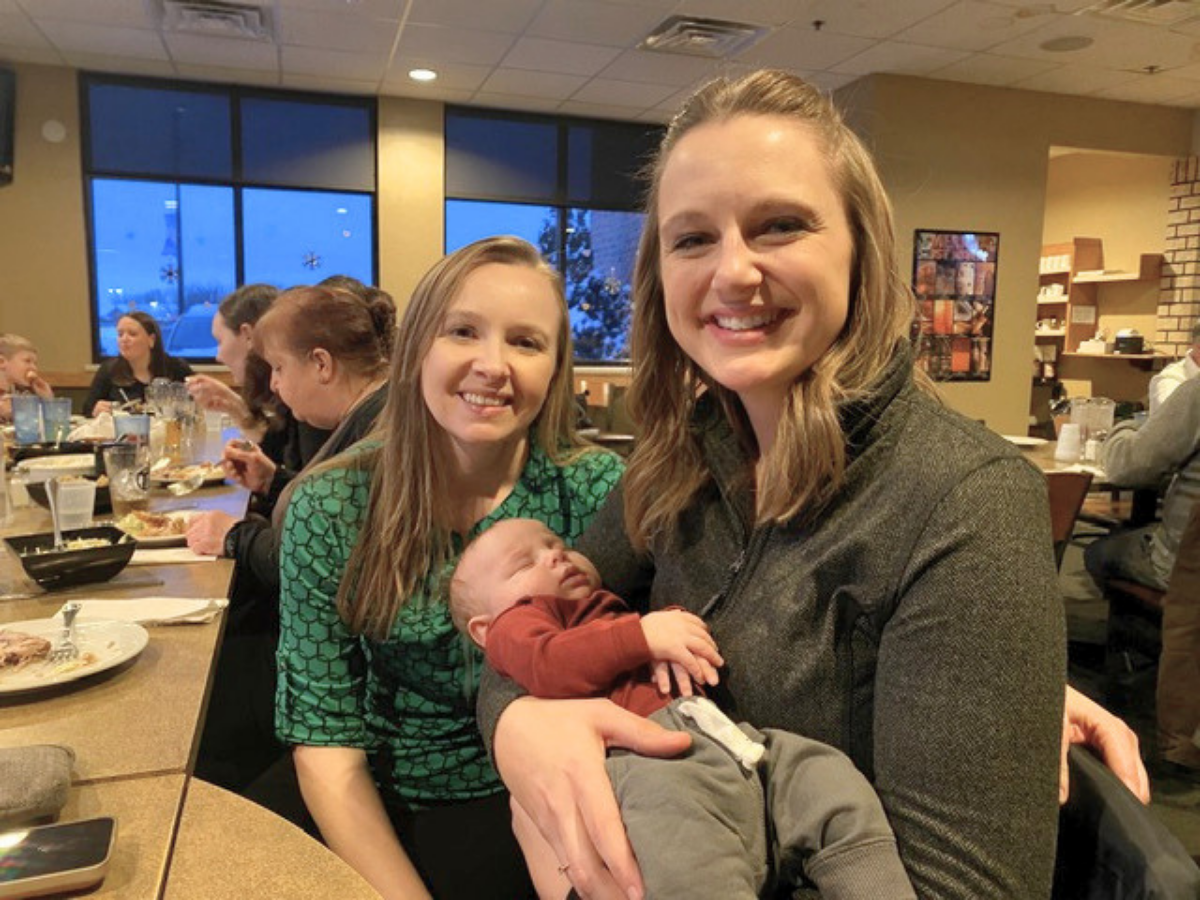 Two smiling women sit at a restaurant table, one holding a sleeping baby, with other diners in the background.