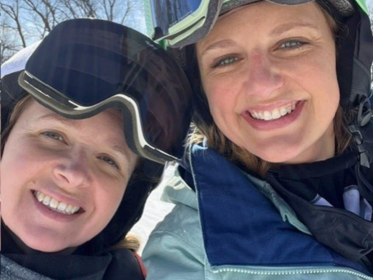 Two women in winter ski gear smiling for a selfie in a snowy outdoor setting, with one wearing black ski goggles on her head.