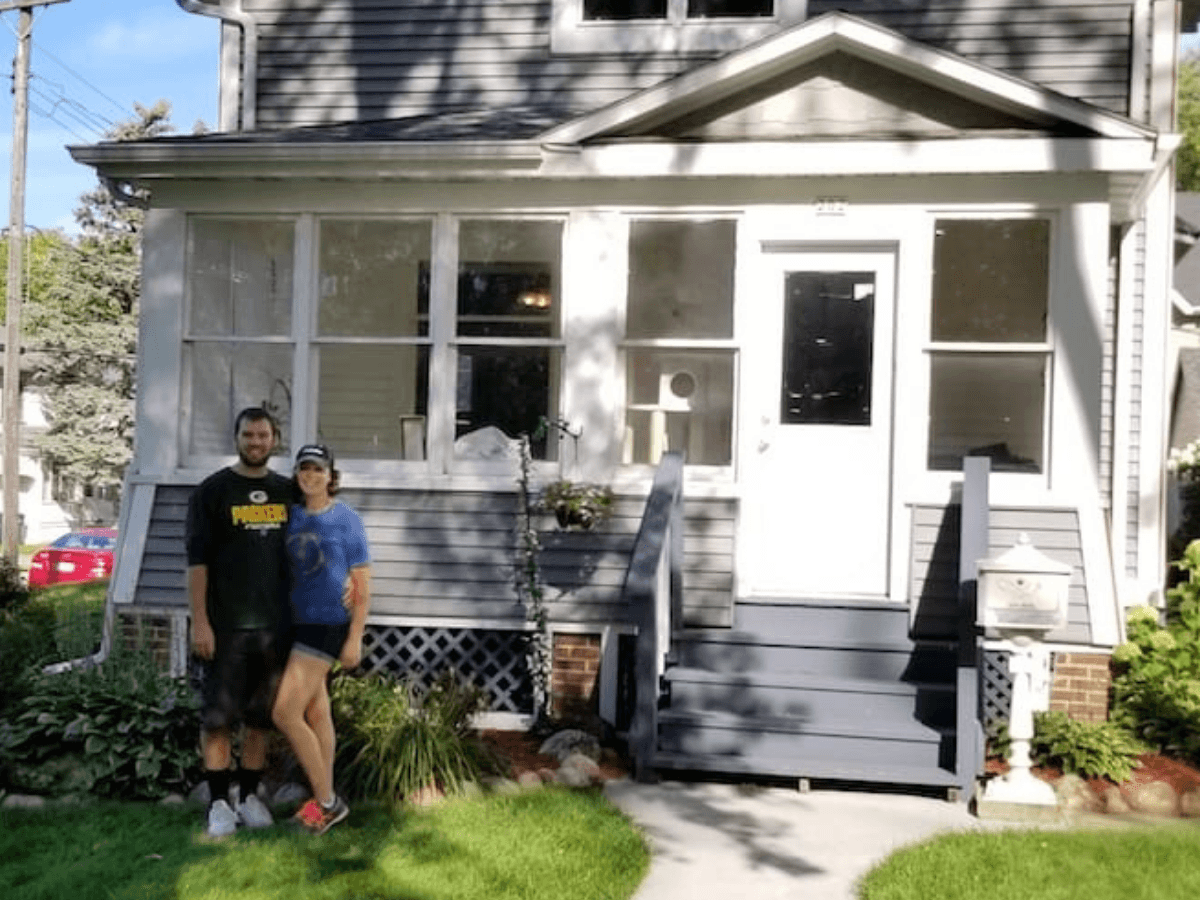 Two people stand on the front lawn in front of a gray house with a small porch and white door.