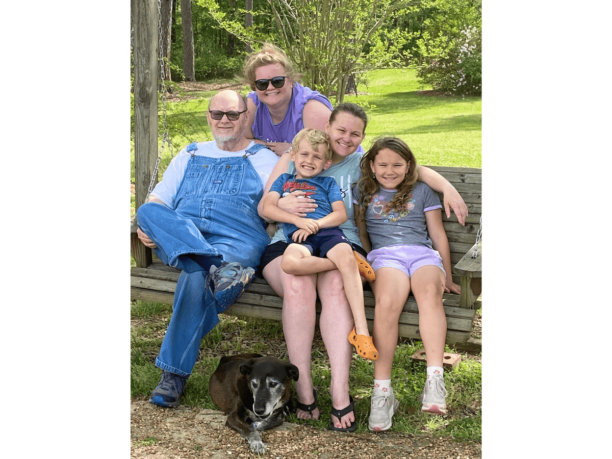 Family sitting on a wooden swing bench in a sunny yard, smiling at the camera with a black-and-white dog resting underneath.