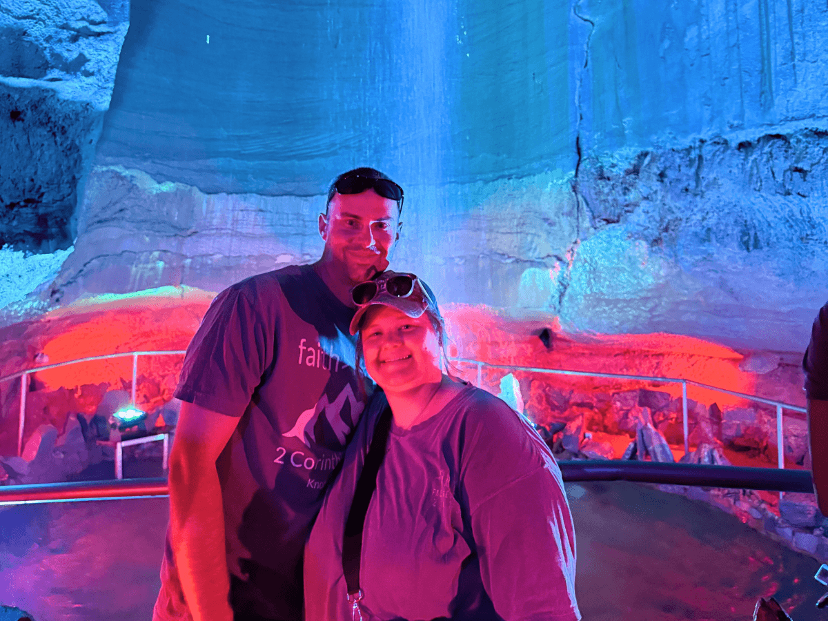 Smiling couple posing in a blue- and red-lit cavern with rocky walls and a railing behind them.