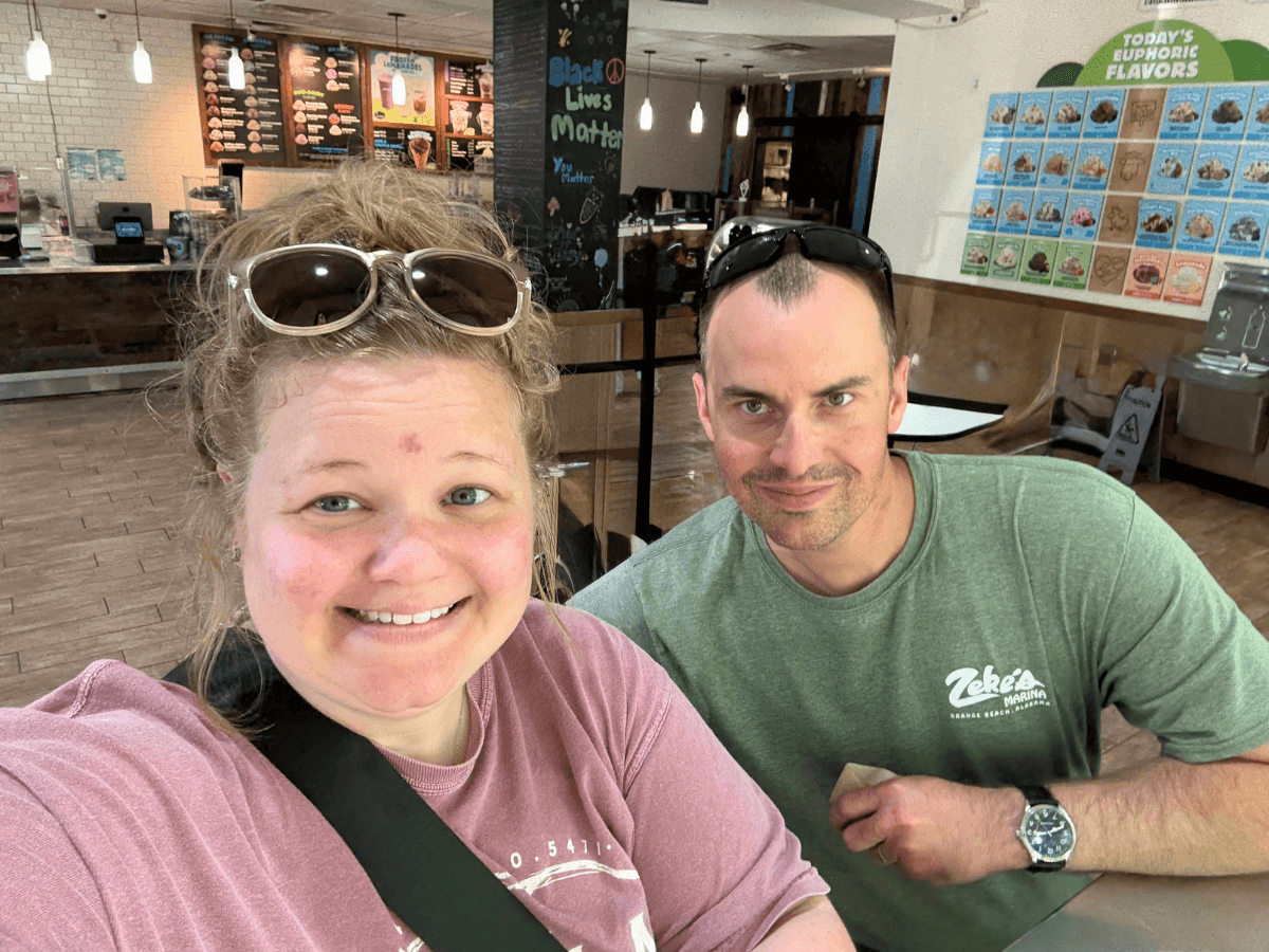 Smiling woman and man take a selfie at a cafe counter, with menu boards and warm hanging lights behind them.