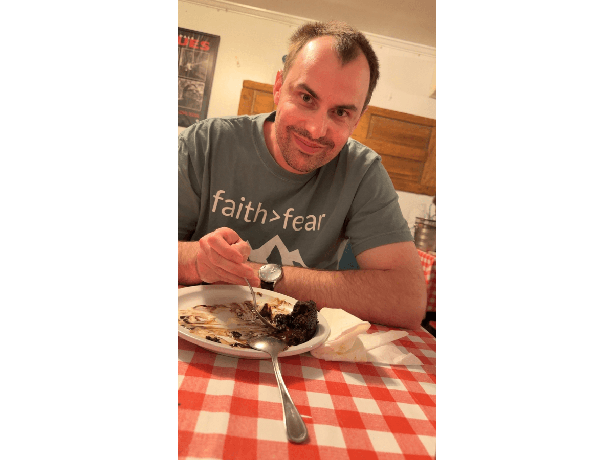 Man in a gray 'faith > fear' shirt smiles while eating chocolate cake at a restaurant table.