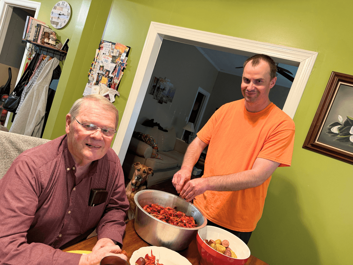 Two men in a green-walled kitchen stand and sit at a table with a large pot of red crawfish; one stirs the pot while the other smiles nearby, with a dog in the doorway.