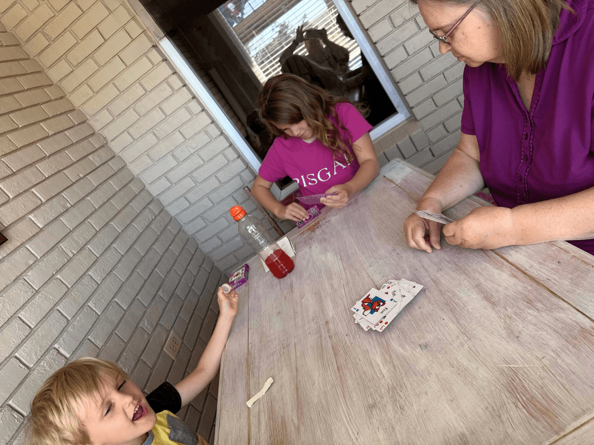 Family of three playing cards at a weathered wooden table on a patio, with a woman counting cards and two kids preparing their hands.