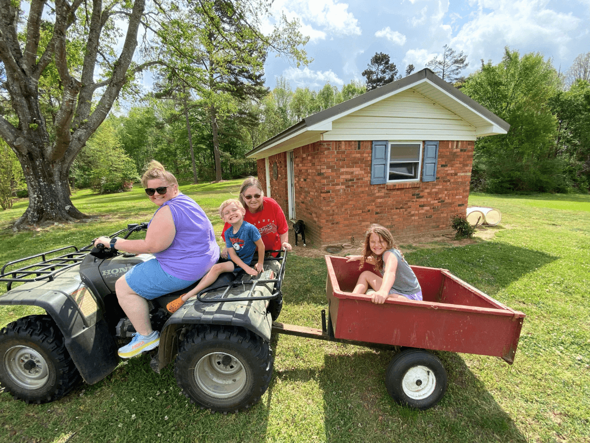 Family outdoors in a yard: woman on an ATV pulling a red wagon with two kids riding beside a brick house.
