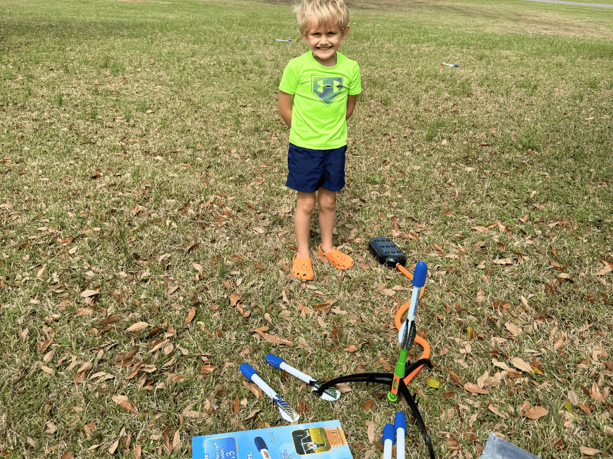 Smiling young boy in a neon green shirt and navy shorts stands on grass beside a partially assembled scooter and scattered toy parts on the ground.
