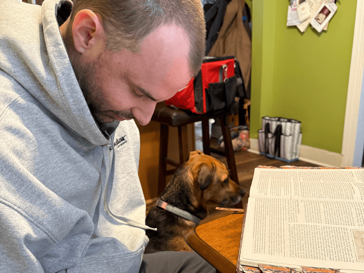 Man in a gray hoodie reads a large open book at a table, with a small dog sitting on the floor behind him in a home setting.