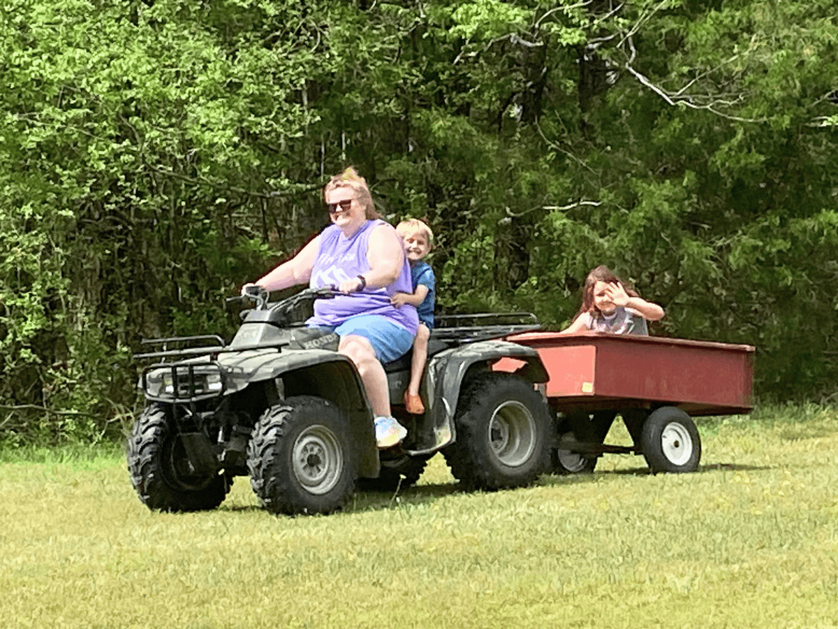 A woman driving an ATV with a child as a passenger, pulling a red trailer holding two kids waving from the back in a grassy, tree-lined field