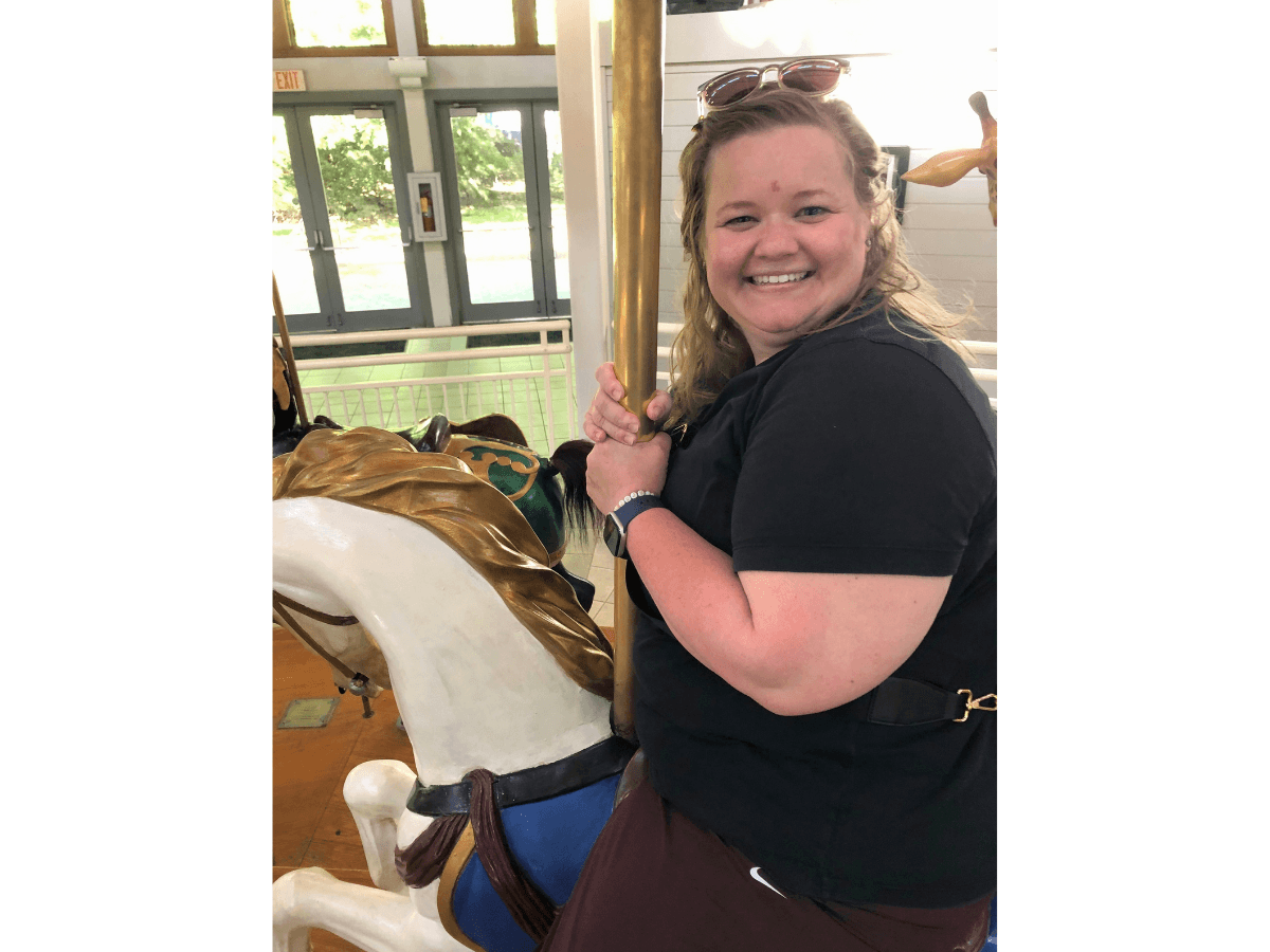 Smiling woman sits on a carousel horse indoors, holding a vertical pole with sunglasses on her head.