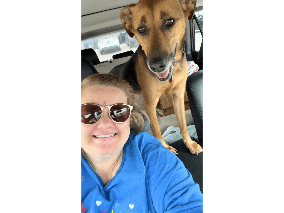 Woman taking a selfie in a car, smiling with a brown dog standing on the back seat beside her, the dog's paws on the seat.