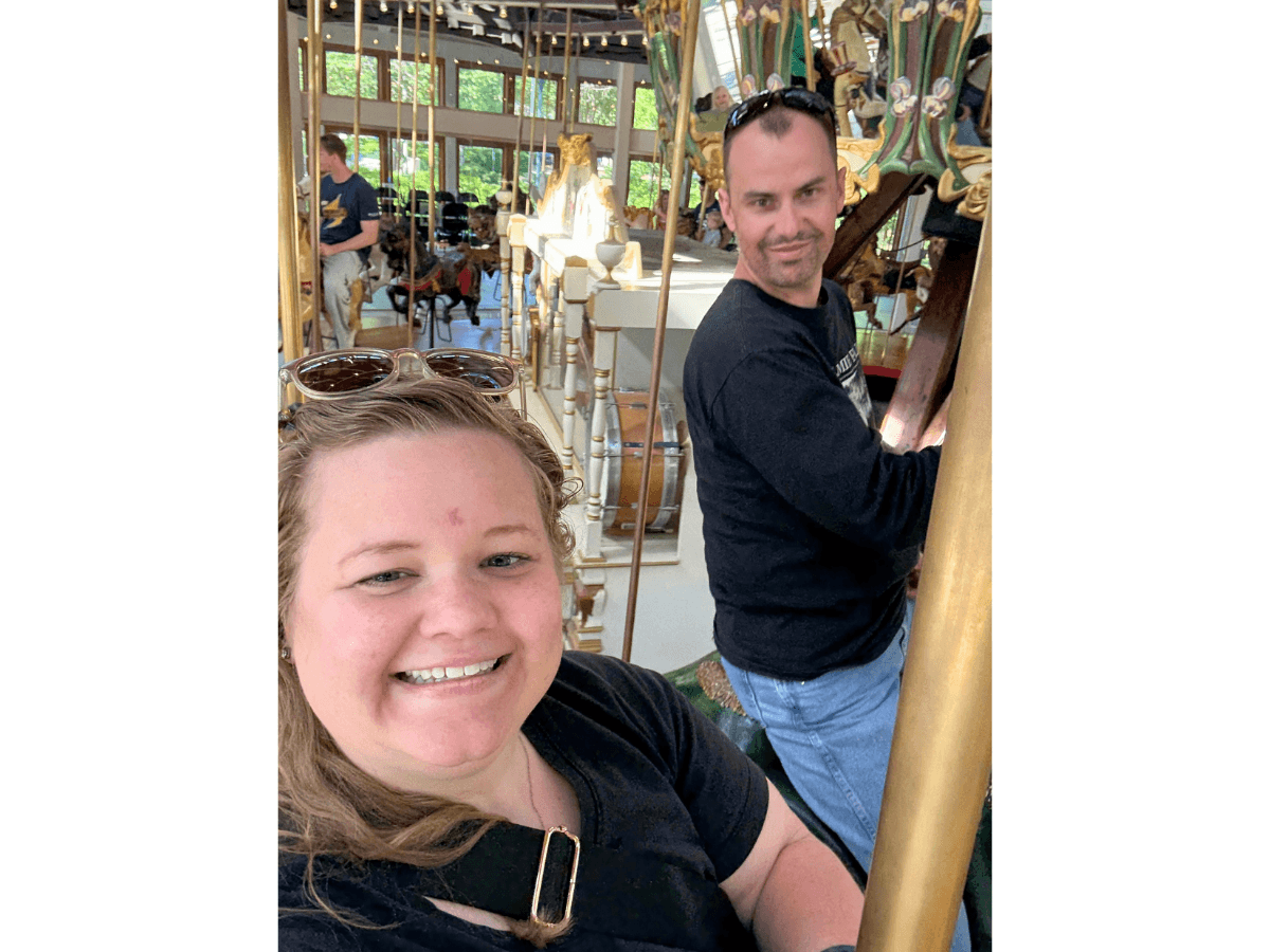 Smiling woman taking a selfie on a carousel, with a man riding a gold pole behind her.