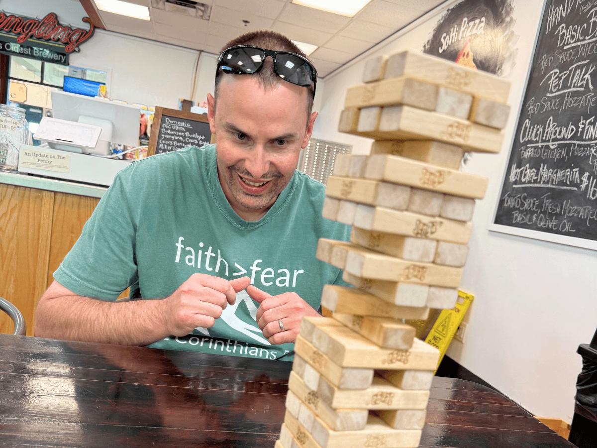 Man wearing sunglasses on his head leans toward a tall wooden Jenga-style block tower on a table, smiling in a shop or cafe setting
