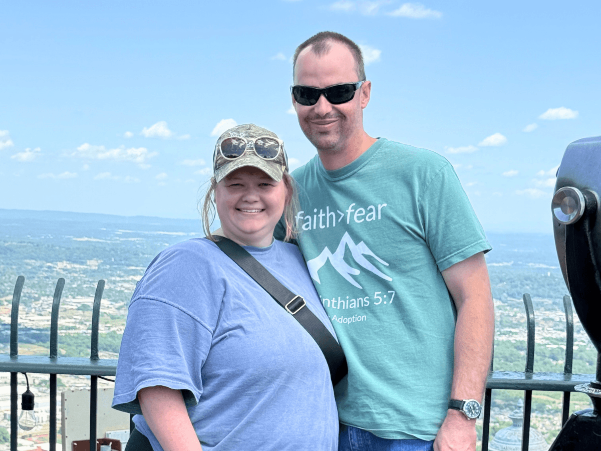 Smiling couple posing at a scenic overlook with a valley and blue sky in the background, railing visible behind them.