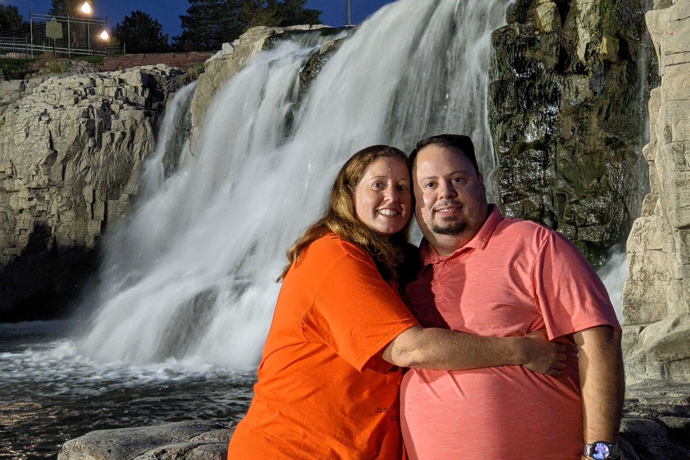Smiling couple hugging in front of a cascading waterfall at dusk or night with rocky surroundings behind them