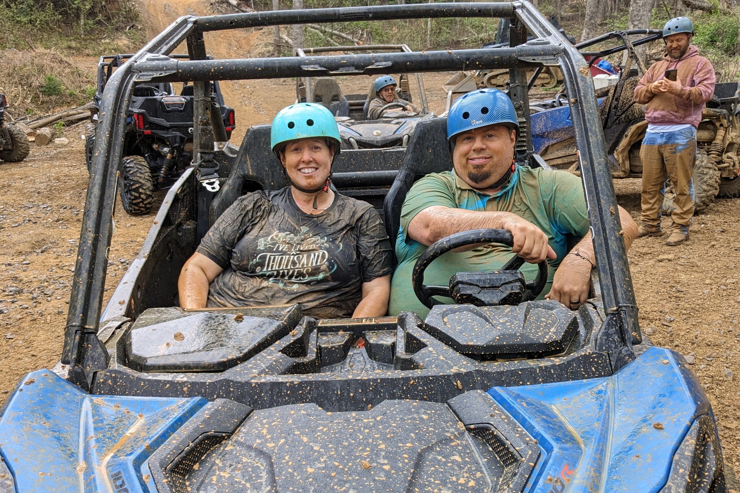 Two people wearing helmets sit in a muddy off-road dune buggy and smile at the camera, with others standing nearby in a dirt trail scene.
