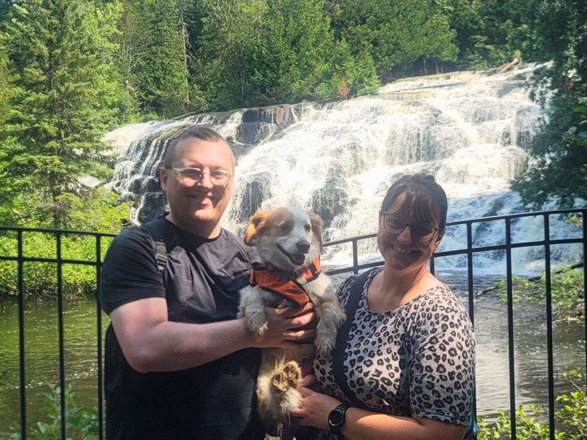 Smiling couple holds a small dog wearing an orange life jacket, with a cascading waterfall and railing behind them.