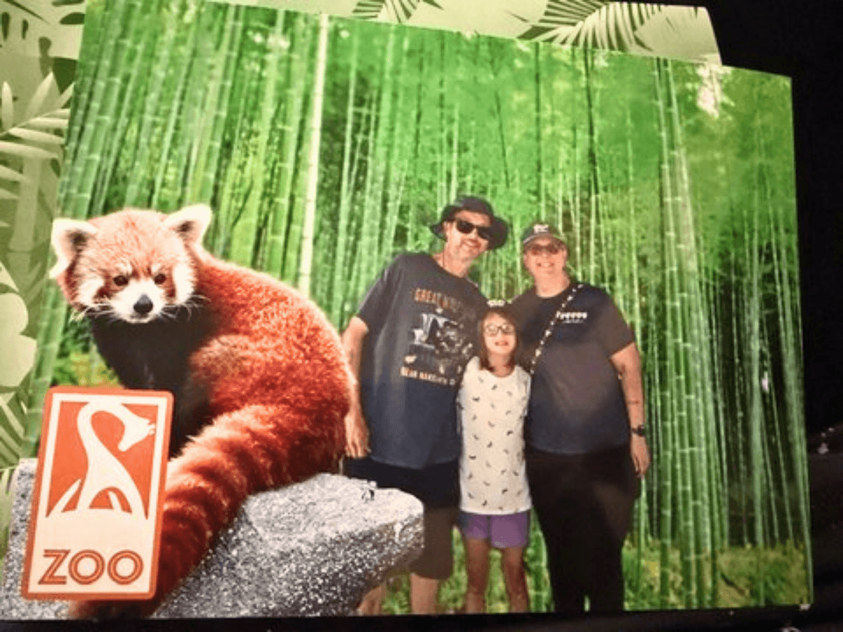 Family of three posing with a life-size red panda statue at a zoo exhibit with a 'Zoo' sign and bamboo backdrop