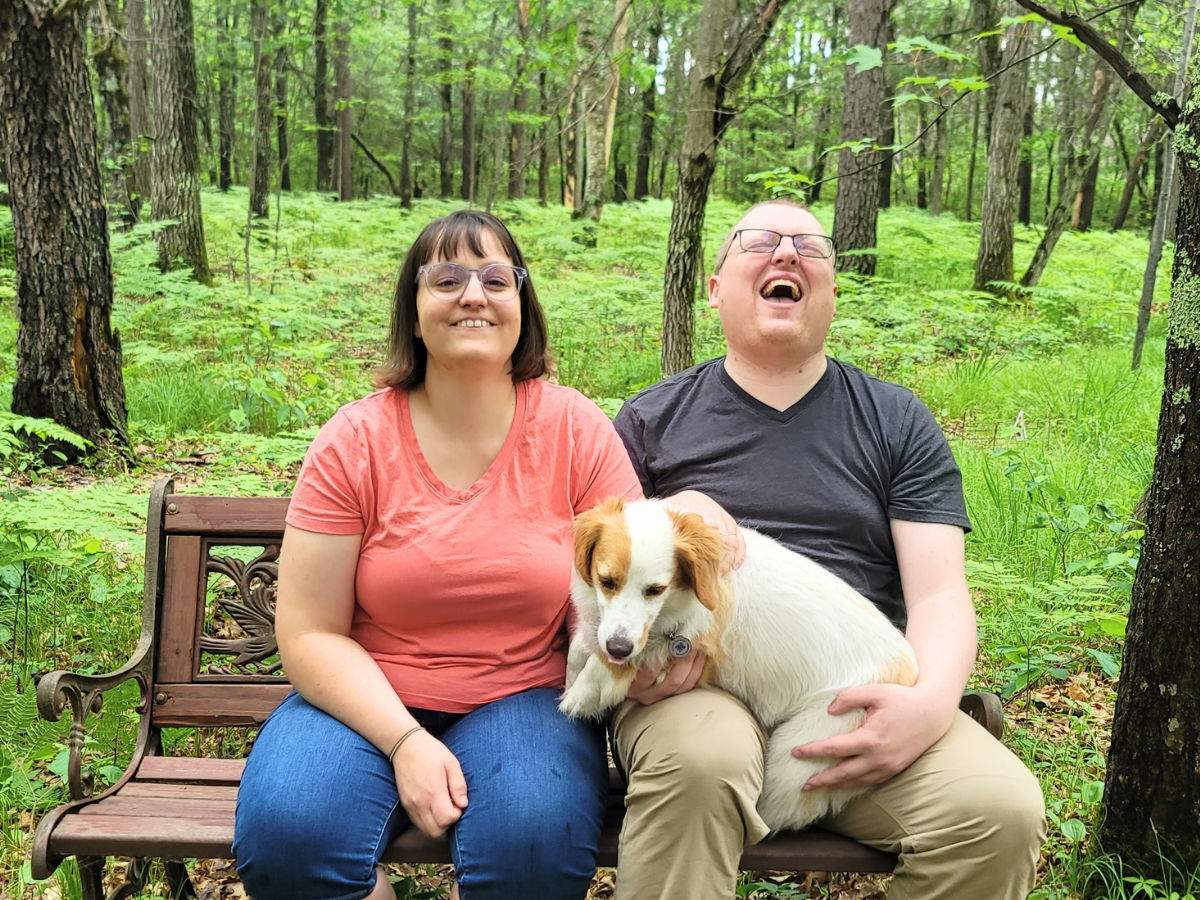 Couple sitting on a wooden bench in a green forest with a small white‑and‑brown dog on the man's lap, the man laughing tall behind them.