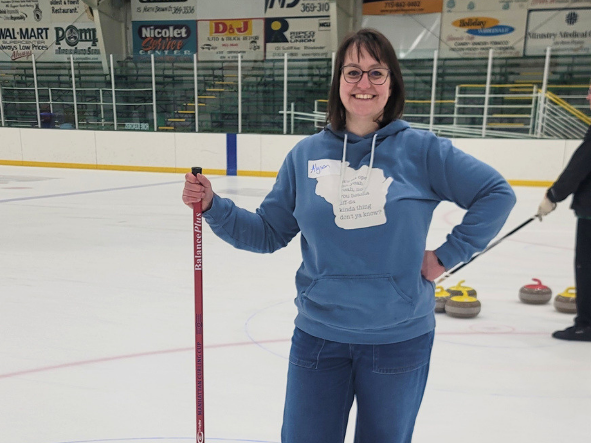 Smiling woman on an indoor ice rink holding a red curling broom, with curling stones visible in the background.