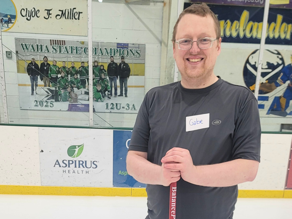 Smiling man in a gray shirt with a name tag 'Gabe' standing on an ice rink holding a hockey stick.