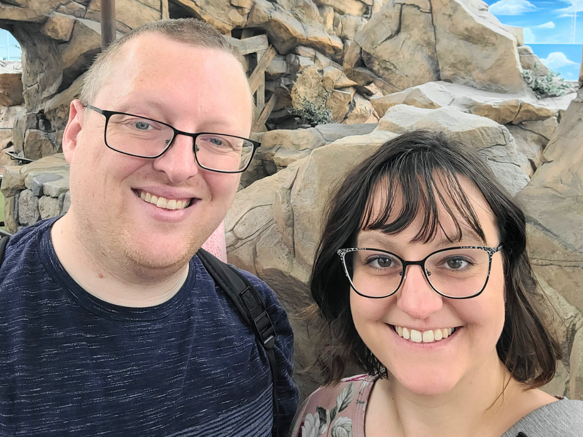 Smiling man and woman wearing glasses pose for a selfie in front of a rock wall backdrop.