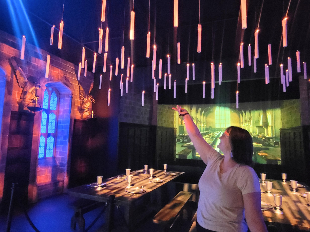 Woman in a white shirt points up at suspended cylindrical lights in a dim, fantasy-style dining hall with a long table set for guests.