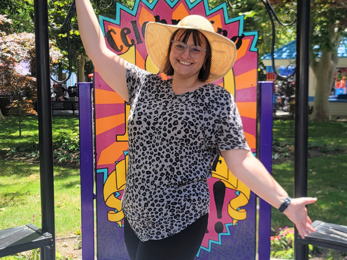 Smiling woman in a leopard-print blouse and wide-brim hat poses with arms raised in front of a colorful mural in a park.