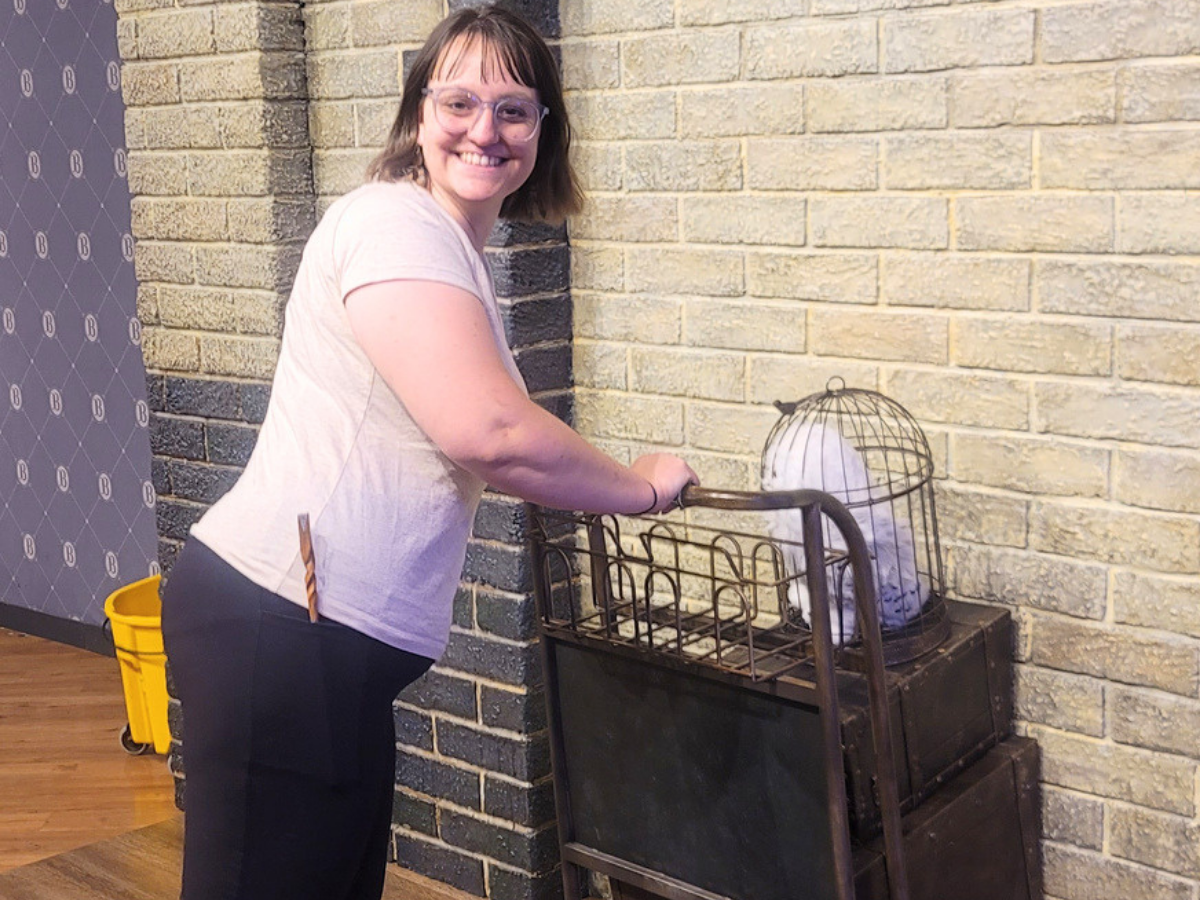 Smiling woman in a pink shirt pushes a wheeled cart with a bird cage on a brick wall background.